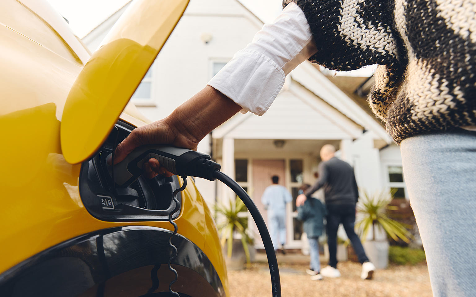 Person plugging a home EV charging cable into a yellow electric vehicle parked outside a residential house.